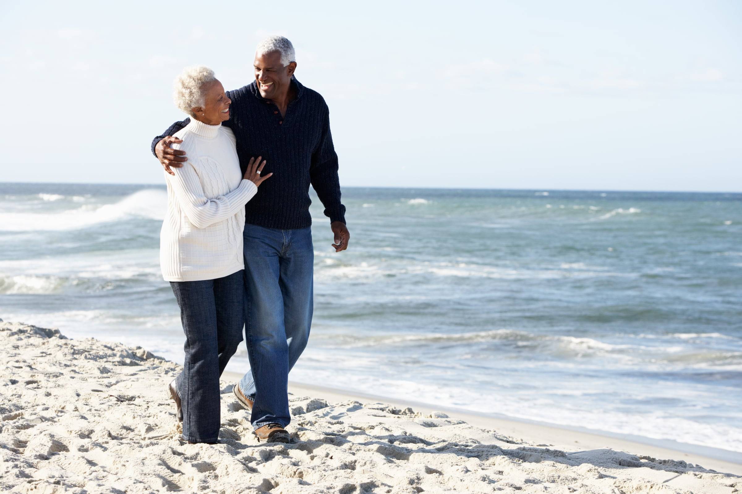 An older couple walks arm in arm along a sandy beach, smiling at each other, with the ocean behind them.