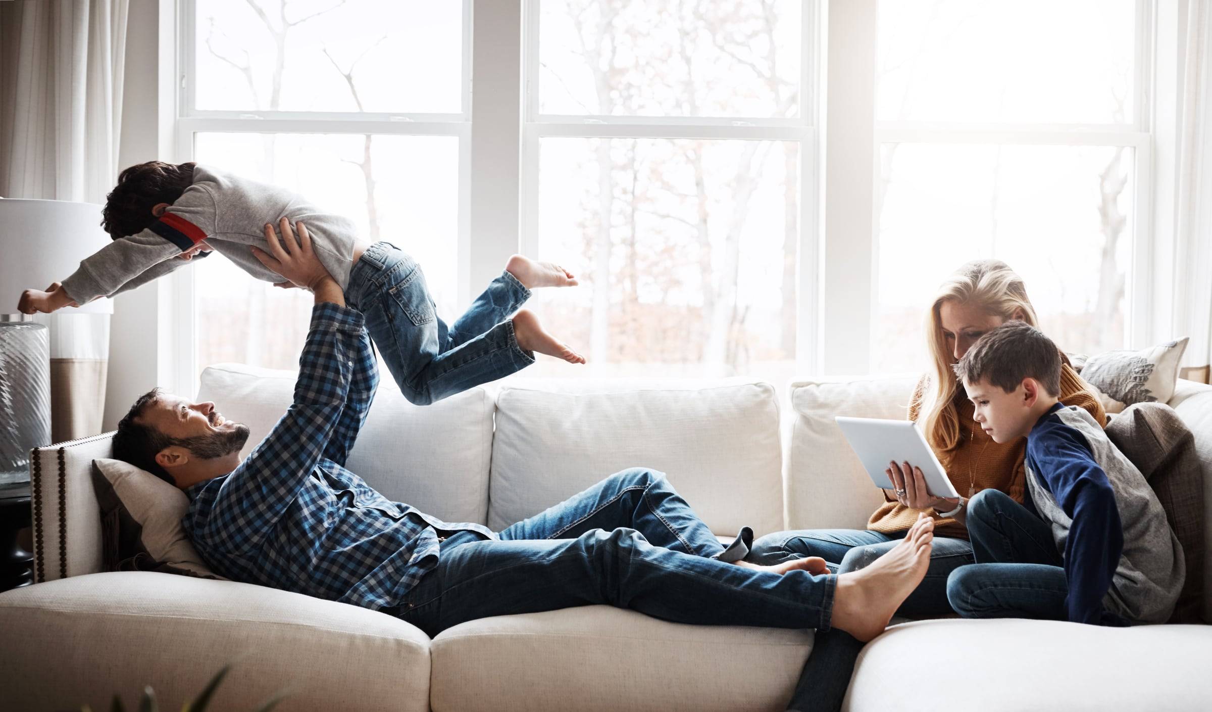 A father on the sofa lifts his young son into the air while the mother and older son look at a tablet beside them.